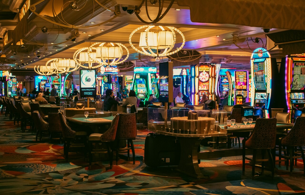 Uma sala de jogos de um cassino físico com pessoas, mesas e máquinas caça-níqueis.